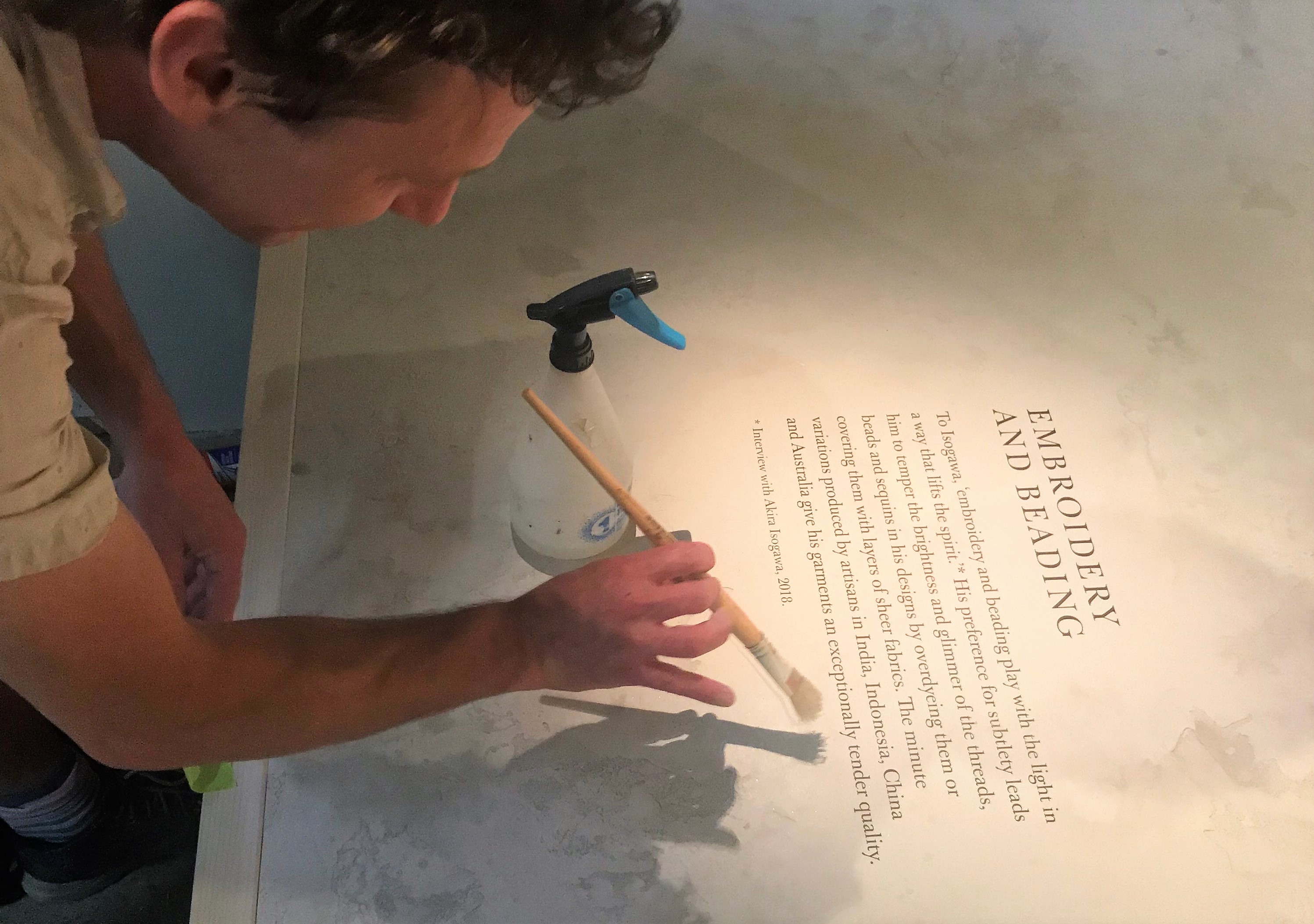 A man with a paintbrush poised over a plasterwork plinth. Written on the plinth is exhibition text with the heading 'embroidering and beading'. There is a spray bottle next to the man's hand.